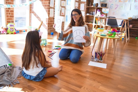 Beautiful pshychologist and toddler doing therapy with emotions emoji around lots of toys at kindergarten