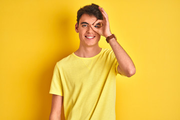 Teenager boy wearing yellow t-shirt over isolated background doing ok gesture with hand smiling, eye looking through fingers with happy face.