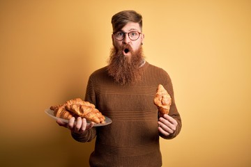 Redhead Irish man with beard eating french croissant pastry over yellow background scared in shock...