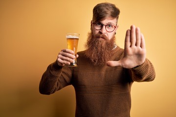 Irish redhead man with beard drinking a glass of refreshing beer over yellow background with open hand doing stop sign with serious and confident expression, defense gesture