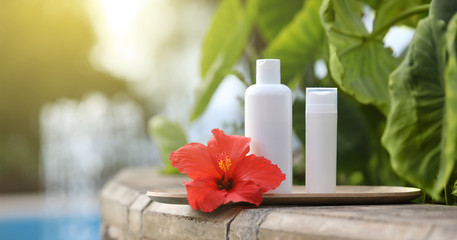 White empty tube of skincare bottle on bamboo plate with red hibiscus over tropical leaves background. Skincare spray tube on blue pool border.