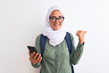 Young Arab student woman wearing hijab using smartphone over isolated background pointing and showing with thumb up to the side with happy face smiling