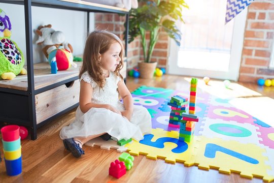 Adorable blonde toddler playing with building blocks toy around lots of toys at kindergarten