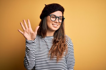 Young beautiful brunette woman wearing french beret and glasses over yellow background showing and pointing up with fingers number five while smiling confident and happy.