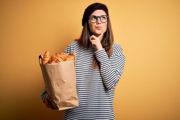 Young beautiful brunette french woman wearing beret holding bag with homemade croissants serious...