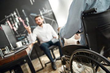 Fototapeta na wymiar Cropped photo of office worker in a wheelchair discussing something with his male colleague while working together in the modern office