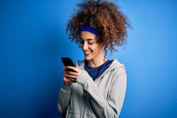 Beautiful sportswoman with curly hair and piercing wearing sportswear using smartphone with a happy face standing and smiling with a confident smile showing teeth