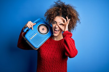Obraz premium Young beautiful woman with curly hair and piercing listening to music using vintage radio with happy face smiling doing ok sign with hand on eye looking through fingers