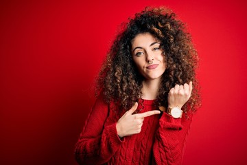 Young beautiful woman with curly hair and piercing wearing casual red sweater In hurry pointing to...