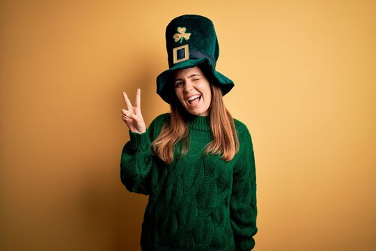 Young beautiful brunette woman wearing green hat on st patricks day celebration smiling with happy face winking at the camera doing victory sign. Number two.