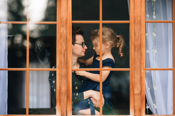 Handsome woman with pretty face hugs a beautiful daughter and looks through big window