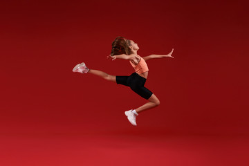 Fototapeta na wymiar Do your best. A cute kid, girl is engaged in sport, she is in motion jumping over in the air. Isolated on red background. Fitness, training, active lifestyle concept. Horizontal shot