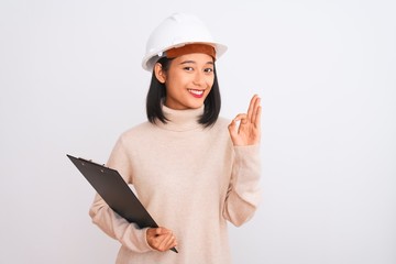 Chinese architect woman wearing helmet holding clipboard over isolated white background doing ok sign with fingers, excellent symbol