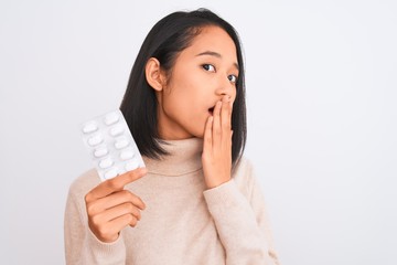Young beautiful chinese woman holding pills standing over isolated white background cover mouth...