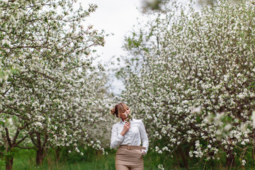 Naklejka na ściany i meble Beautiful young blonde woman in white shirt posing under apple tree in blossom in Spring garden