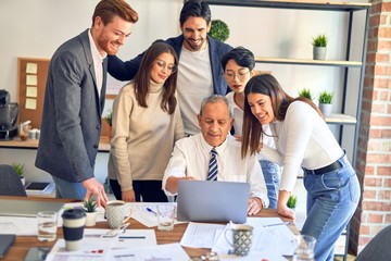 Fototapeta premium Group of business workers smiling happy and confident. One of them sitting and partners standing around. Working together with smile on face looking at the laptop at the office