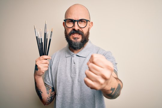 Handsome bald artist man with beard and tattoo painting using painter brushes annoyed and frustrated shouting with anger, crazy and yelling with raised hand, anger concept