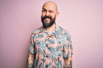 Handsome bald man with beard and tattoo wearing casual floral shirt over pink background winking looking at the camera with sexy expression, cheerful and happy face.