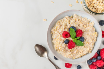 Bowl of oatmeal porridge with fresh berries and mint served with spoon for healthy diet breakfast on white table. top view