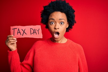 Young African American afro woman with curly hair holding paper with tax day message scared in...