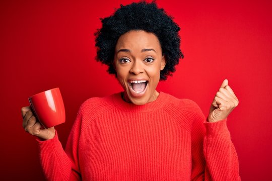 African American afro woman with curly hair drinking cup of coffee over red background screaming proud and celebrating victory and success very excited, cheering emotion