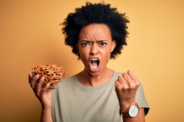 Young African American afro woman with curly hair holding bowl with baked German pretzels annoyed and frustrated shouting with anger, crazy and yelling with raised hand, anger concept