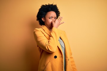 Young beautiful African American afro businesswoman with curly hair wearing yellow jacket smelling something stinky and disgusting, intolerable smell, holding breath with fingers on nose. Bad smell