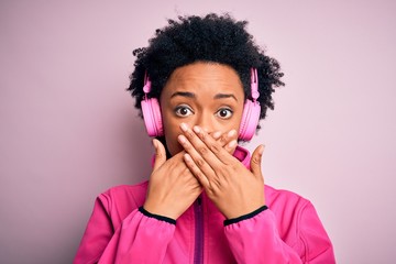 Young African American afro woman with curly hair listening to music using pink headphones shocked covering mouth with hands for mistake. Secret concept.