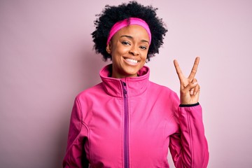 Young beautiful African American afro sportswoman with curly hair wearing pink sportswear smiling with happy face winking at the camera doing victory sign. Number two.