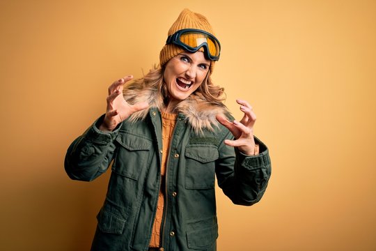 Middle age beautiful blonde skier woman wearing snow sportwear and ski goggles Shouting frustrated with rage, hands trying to strangle, yelling mad
