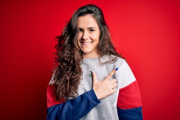 Young beautiful woman with curly hair wearing casual sweatshirt over isolated red background cheerful with a smile on face pointing with hand and finger up to the side