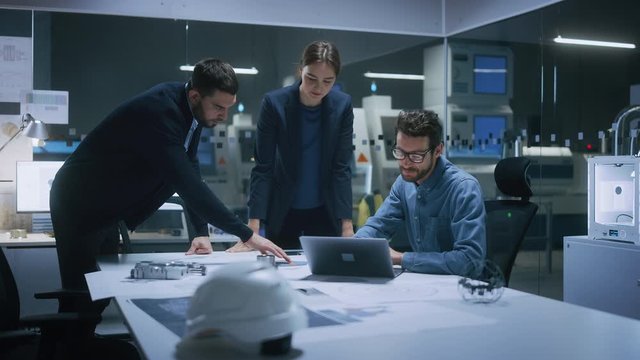 Modern Factory Office Meeting Room: Diverse Team of Engineers, Managers Talking at Conference Table, Look at Blueprints, Inspect Mechanism, Use Laptop. High-Tech Facility with CNC Machines