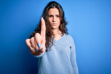 Young beautiful woman with curly hair wearing blue casual sweater over isolated background Pointing with finger up and angry expression, showing no gesture