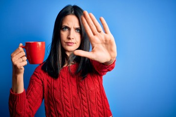 Young woman with blue eyes drinking cup of coffee standing over isolated background with open hand doing stop sign with serious and confident expression, defense gesture