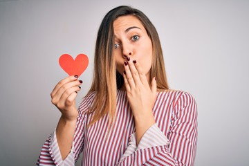 Young beautiful blonde romantic woman holding red paper heart over white background cover mouth with hand shocked with shame for mistake, expression of fear, scared in silence, secret concept