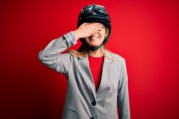 Young beautiful blonde motorcyclist woman wearing motorcycle helmet over red background smiling and laughing with hand on face covering eyes for surprise. Blind concept.
