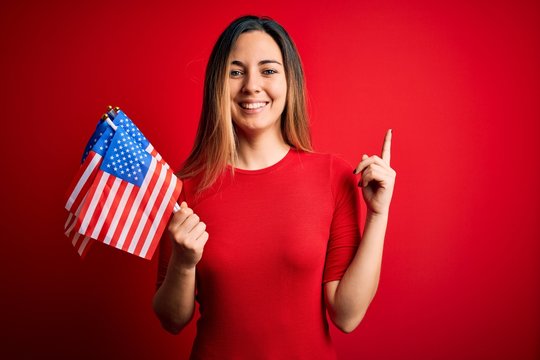 Young beautiful blonde patriotic woman with blue eyes holding united states flags surprised with an idea or question pointing finger with happy face, number one