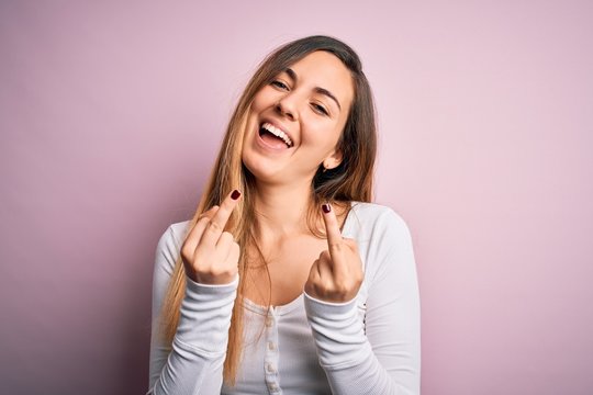 Young beautiful blonde woman with blue eyes wearing white t-shirt over pink background Showing middle finger doing fuck you bad expression, provocation and rude attitude. Screaming excited