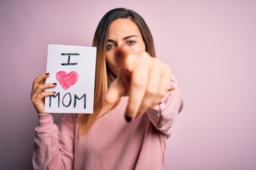 Young beautiful woman holding paper with love mom message celebrating mothers day pointing with finger to the camera and to you, hand sign, positive and confident gesture from the front