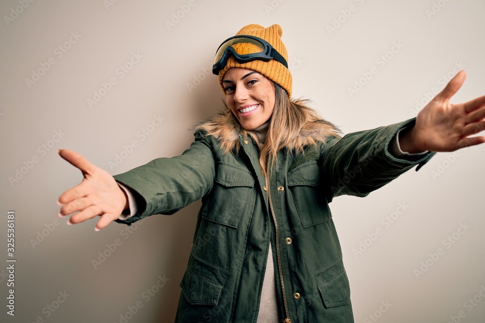 Canvas Prints Young brunette skier woman wearing snow clothes and ski goggles over white background looking at the camera smiling with open arms for hug. Cheerful expression embracing happiness.