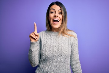 Young beautiful woman wearing casual sweater standing over isolated purple background pointing finger up with successful idea. Exited and happy. Number one.