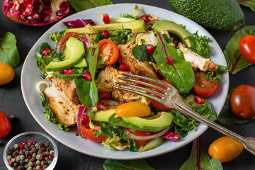 Grilled chicken breast and avocado salad with tomatoes and pomegranate in a plate with fork on dark...
