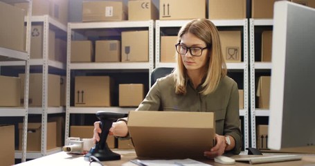 Beautiful Caucasian woman post worker sitting at desk and working at computer in postal delivery store. Female scanning parcel and registering it in system in post office.
