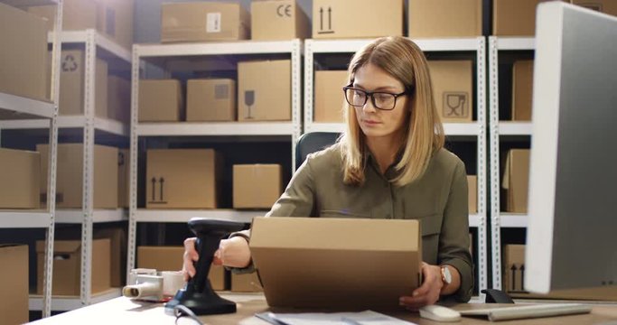 Beautiful Caucasian woman post worker sitting at desk and working at computer in postal delivery store. Female scanning parcel and registering it in system in post office.