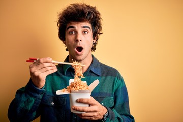 Young handsome man eating noodles standing over isolated yellow background scared in shock with a...