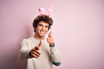 Young handsome man holding easter rabbit ears standing over isolated pink background pointing fingers to camera with happy and funny face. Good energy and vibes.
