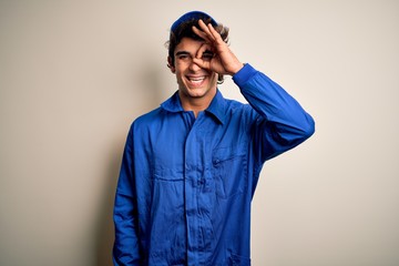 Young mechanic man wearing blue cap and uniform standing over isolated white background doing ok gesture with hand smiling, eye looking through fingers with happy face.