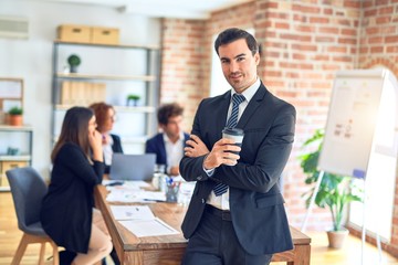 Group of business workers smiling happy and confident working together in a meeting. One of them, standing with smile on face looking at camera drinking coffee at the office.