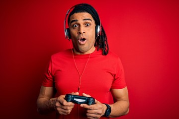 African american man with dreadlocks playing video game using joystick and headphones scared in shock with a surprise face, afraid and excited with fear expression