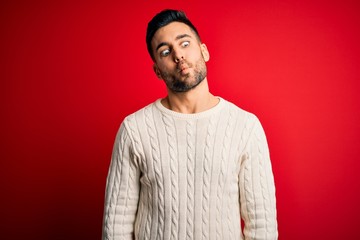 Young handsome man wearing casual white sweater standing over isolated red background making fish face with lips, crazy and comical gesture. Funny expression.